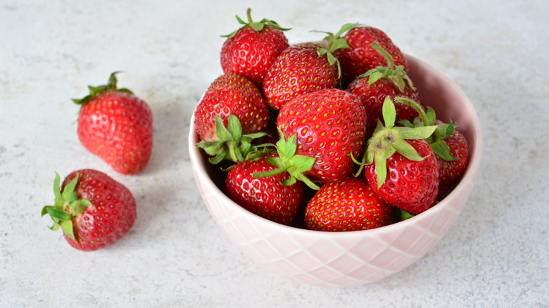 A pink bowl full of fresh strawberries on a marble table with two strawberries next to it