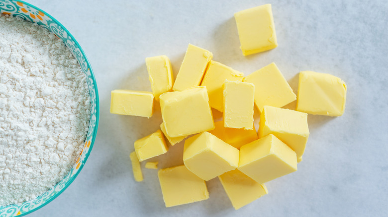 Cold butter cubes on a marble table next to a bowl of flour