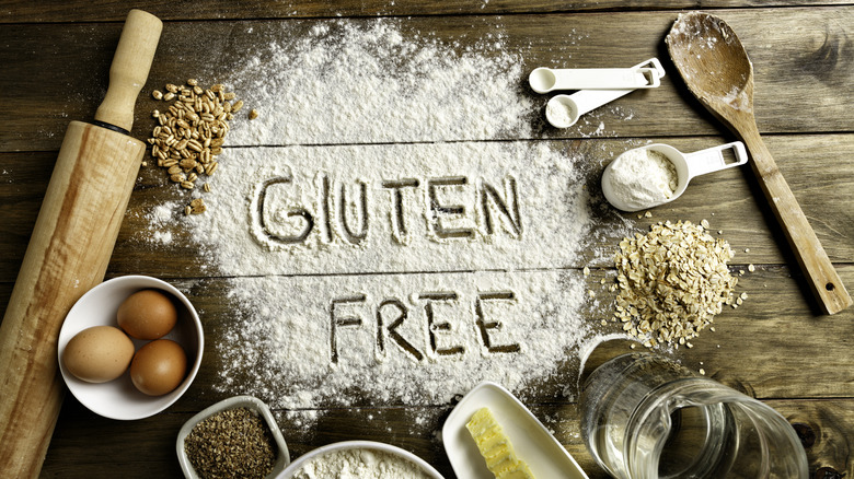 Gluten free is written in flour on a wooden tabletop surrounded by grains and making supplies