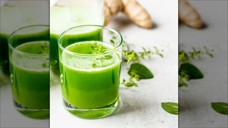 Close-up of a glass of Martha Stewart's green juice with extra in a pitcher, sprigs of greens, and ginger blurred in the background