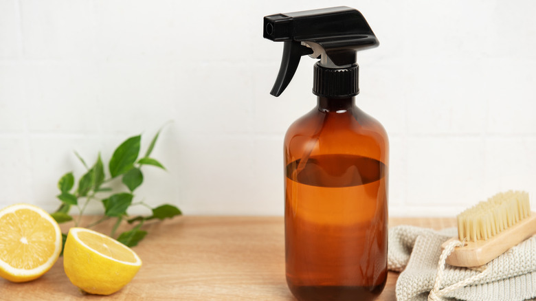 A brown glass spray bottle on a wooden surface with other natural cleaning products and a cut lemon