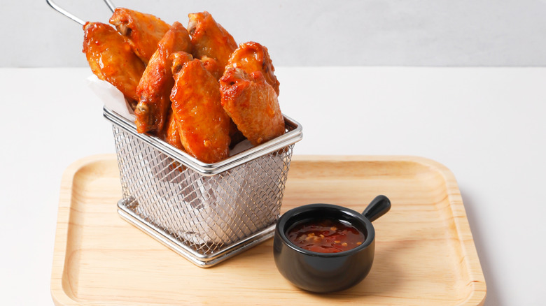 A basket full of chicken wings with sauce on the side, all on a wooden plate