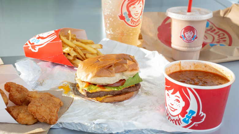 An assortment of Wendy's items, including a Dave's single cheeseburger, chicken nuggets, chili, fries, soda, and a Frosty against a gray background.