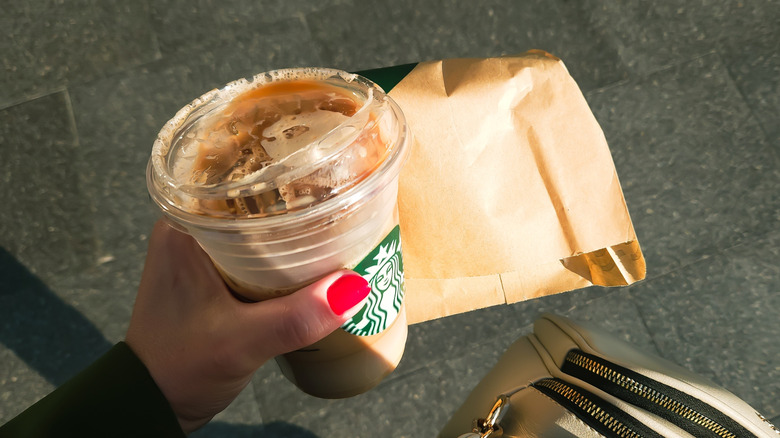 A hand with red-painted nails, holding a Starbucks iced coffee and package of food