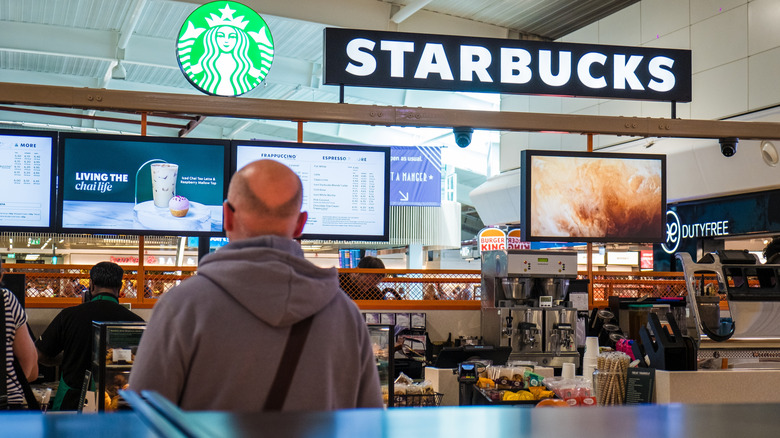 Man standing in front of an airport Starbucks, with its menu and baristas in the background
