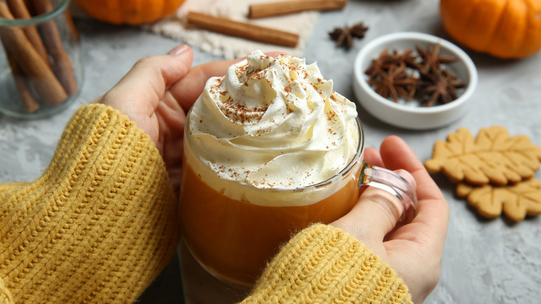 A pair of hands hold aglass mug of pumpkin spice latte with whipped cream