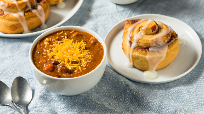 White bowl of chili with cinnamon roll on the side on a white plate