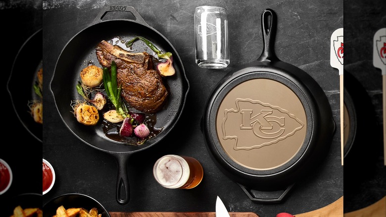 A cast iron skillet cooking a steak with a second skillet overturned, showing the Kansas City Chiefs logo embossed on the bottom