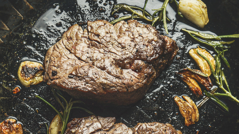 A cooked steak on a black pan with garlic and herbs nearby