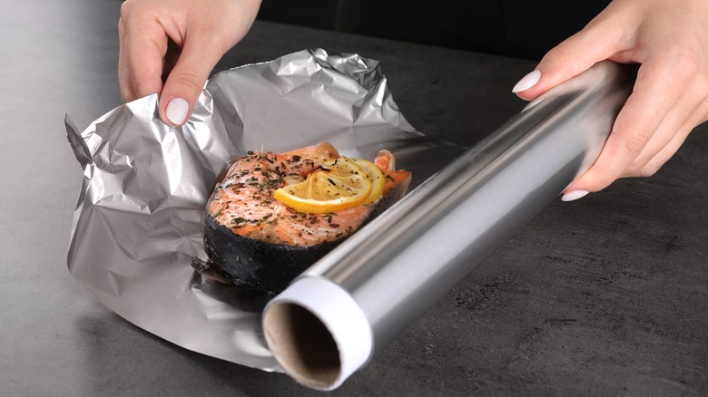 a person's hands wrapping a piece of salmon topped with lemon in aluminum foil