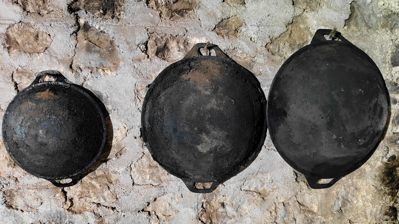 Three different sized cast iron woks with round bottoms hanging on a stone wall