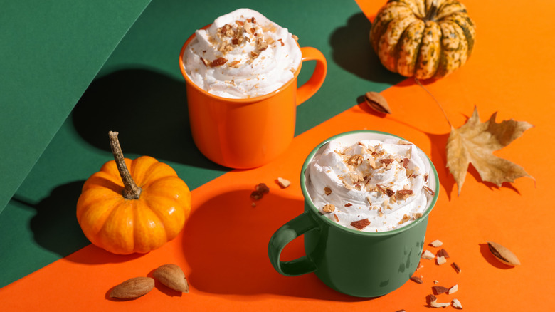 An orange mug and green mug full of coffee and whipped cream next to two pumpkins and leaves, on top of an orange and green background
