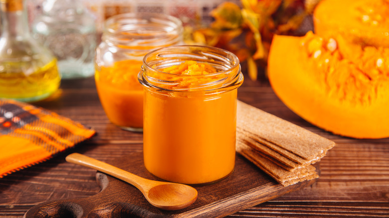 A glass container of pumpkin puree next to a wooden spoon; sliced pumpkins can be seen in the background