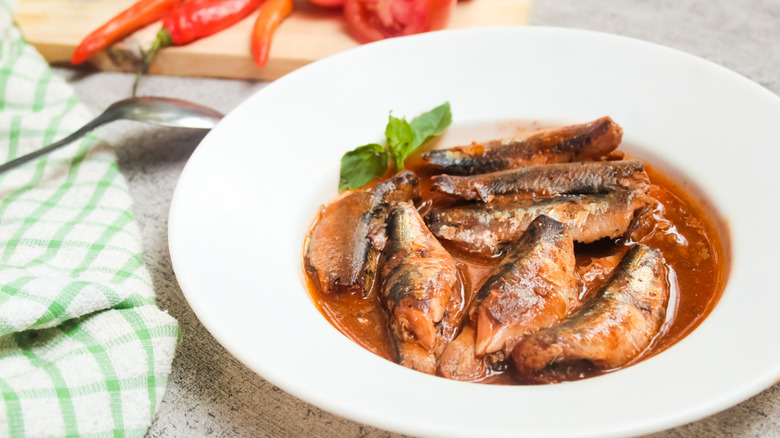 Canned sardine filets in a fresh brownish-red sauce, with tomatoes and chilis in the background