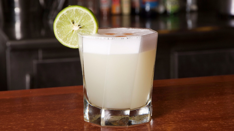 A pisco sour in a rocks glass, garnished with a lemon slice. The murky yellow drink has a foamy top, dotted with three dashes of angostura bitters