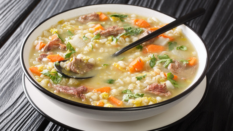 bowl of scotch broth with meat, barley, vegetables, with a spoon against dark background