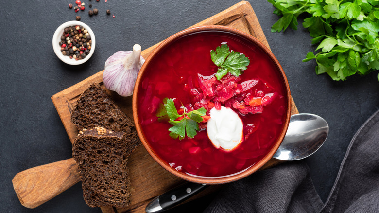 overhead shot of a bowl of red borscht, next to slices of dark brown bread, garlic, peppercorns