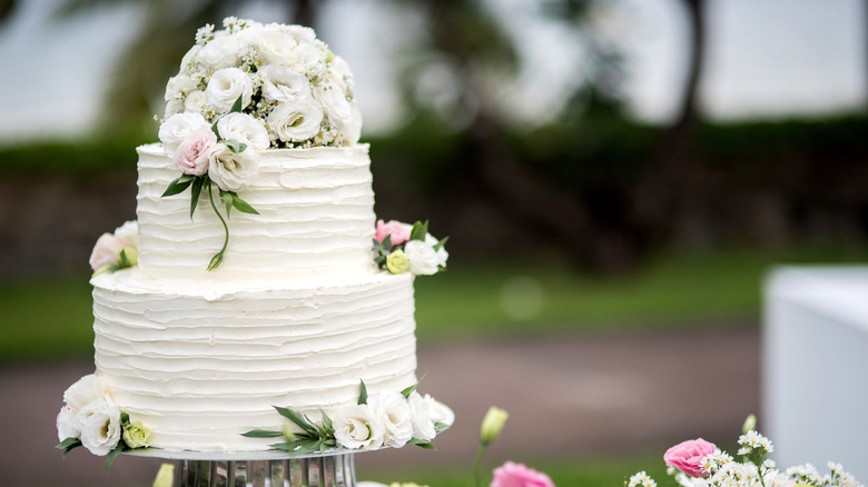 A white, tiered wedding cake decorated with flowers sits on a stand