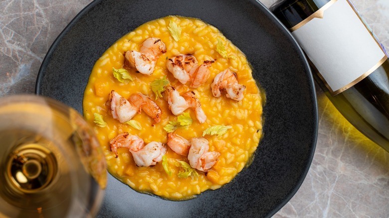 A top-down view of a creamy lobster risotto in a black bowl next to a bottle and glass of wine