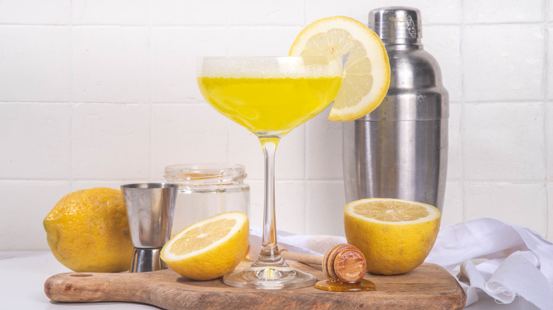 A bright yellow bee's knees cocktail with slices of lemon, honey, and an alcohol shaker beside it