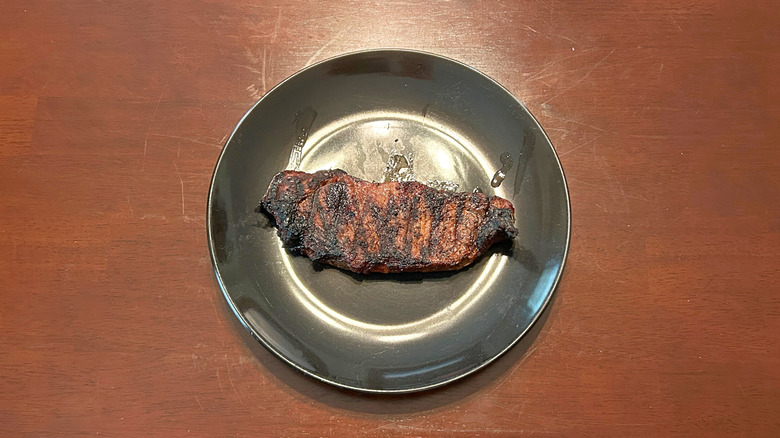 A 12-ounce New York Strip steak with extensive charring and trails of grease on a black plate