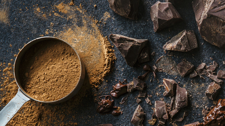 Measuring cup with cocoa powder on a table with chocolate chunks and spilled cocoa powder.