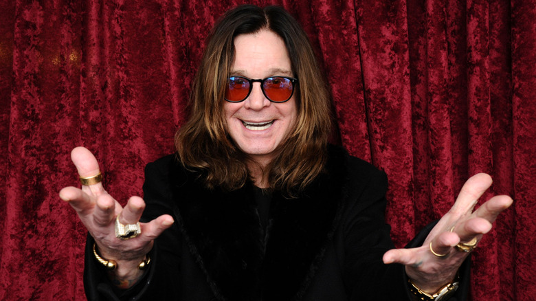 Ozzy Osbourne in front of a red curtain, gesturing to the camera
