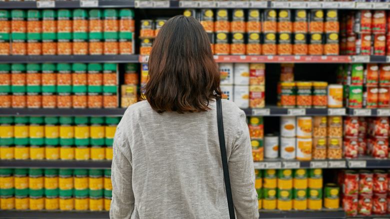 A woman stands in front of a huge selection of canned foods in a supermarket