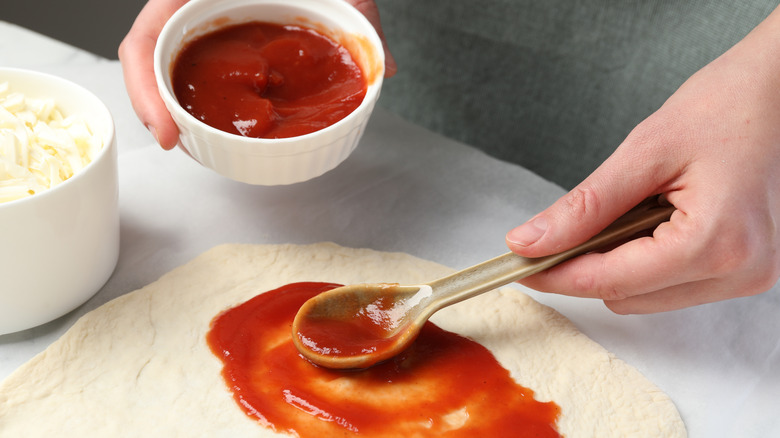 Spreading sauce on pizza dough with a spoon while holding a ramekin of sauce