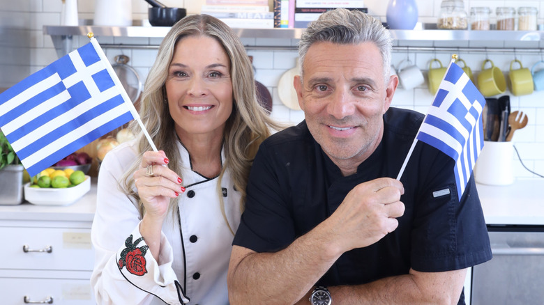 Celebrity chefs Cat Cora and John DeLucie waving Greek flags in a kitchen