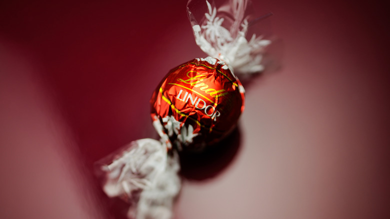 A single Lindt Lindor truffle wrapped in golden orange foil against a maroon backdrop.