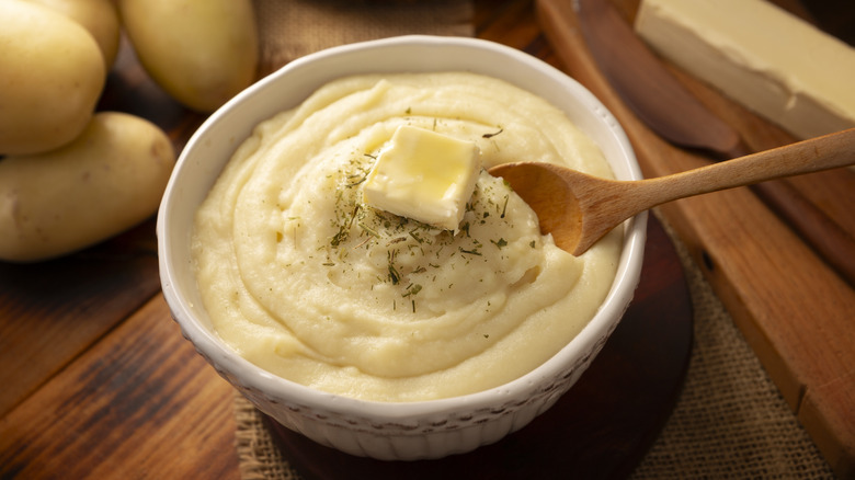 A white bowl of mashed potatoes topped with seasoning and butter with a wooden spoon, in front of potatoes and a stick of butter