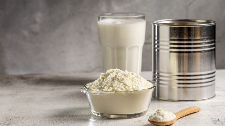 Milk powder in a bowl, and a wooden spoon, a glass of milk, and a can behind them.