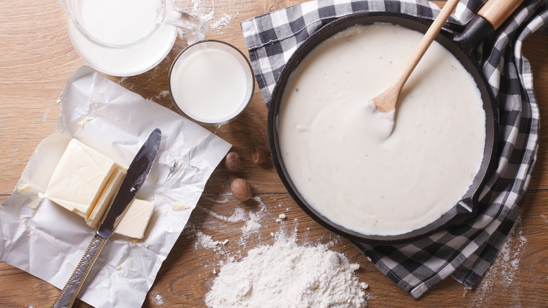 Freshly made béchamel sauce in a pan with a wooden spoon, next to a pitcher and glass of milk, butter on foil, and a pile of milk powder.