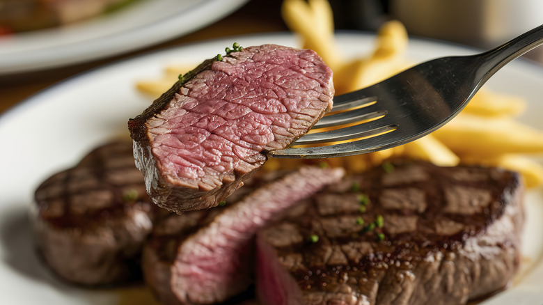 Fork holding piece of medium-rare steak in front of steak and fries