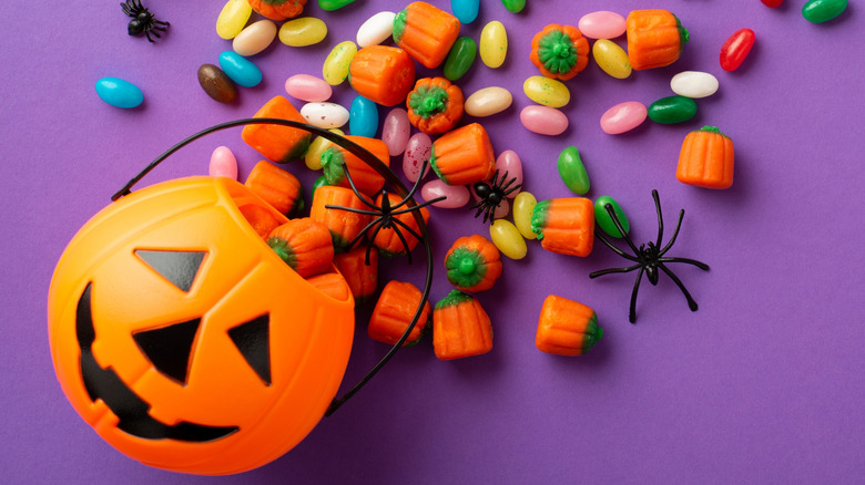 Halloween candy and plastic spiders spilling out of a pumpkin bucket on a purple background