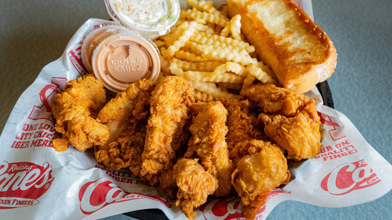 A combo meal from Raising Cane's, with its chicken fingers displayed prominently at the bottom, along with fries, Texas toast, two tubs of Cane's Sauce, and a tub of coleslaw