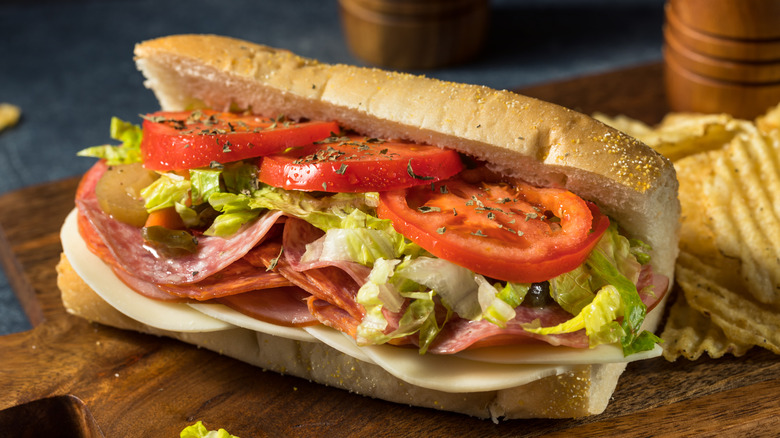 A sub sandwich with meat, cheese, and salad next to potato chips