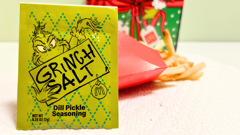 Close up of a Grinch Salt dill pickle seasoning packet with a slightly blurred background consisting of McDonalds fries