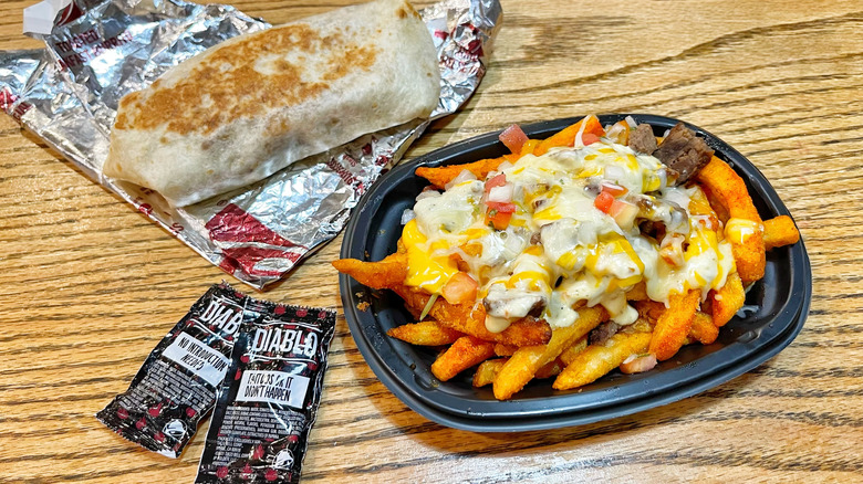 A burrito place on its foil and nacho fries with hot sauce packets on a wood table