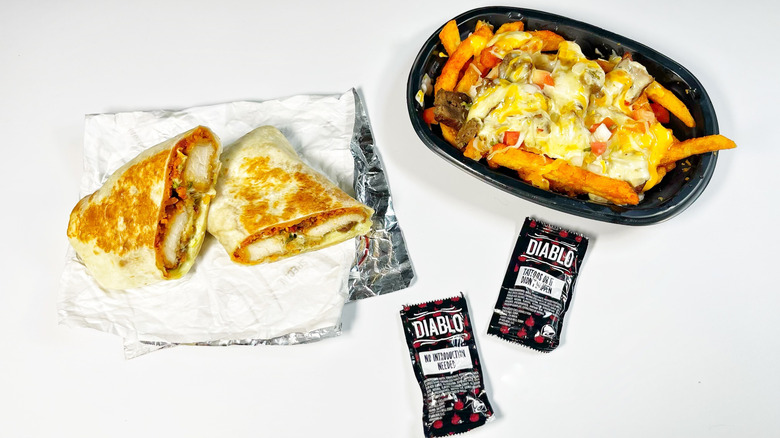 A Taco Bell burrito slice in half on top of paper next to fries and hot sauce packets