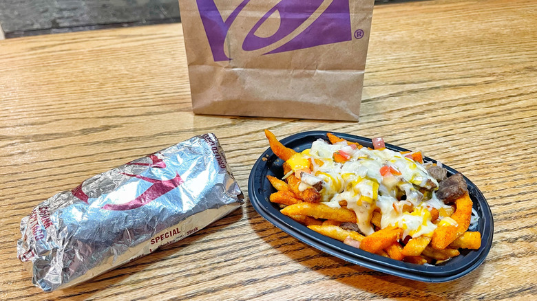 A foil-wrapped burrito and nacho fried on a wooden tabletop next to a Taco Bell bag