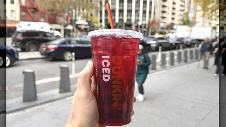 A closeup of a hand holding a Dunkin' Berry Sangria Dunkin' Refresher on a city street