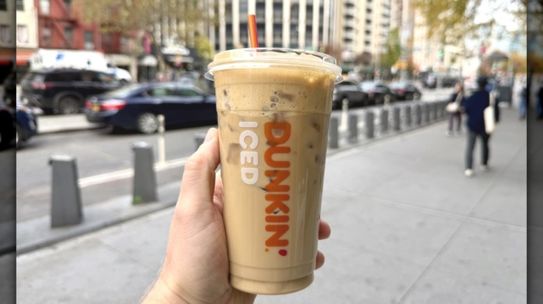 A closeup of a hand holding a Dunkin' Cookie Butter Cloud Latte on a city street