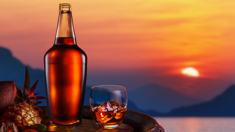 A bottle and glass of dark rum on a barrel with tropical fruit and a sunset background