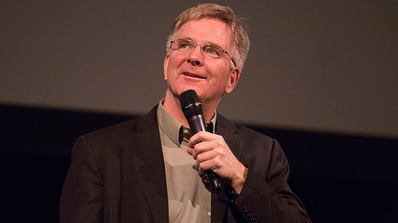 Rick Steves smiling while talking into a microphone against a dark background