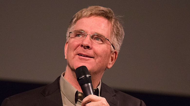 Travel writer Rick Steves speaking into a microphone wearing a suit and glasses