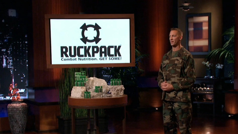 A man in green army camo stands next to a table of green energy shot bottles, with the logo for "RuckPack Combat Nutrition. GET SOME!"
