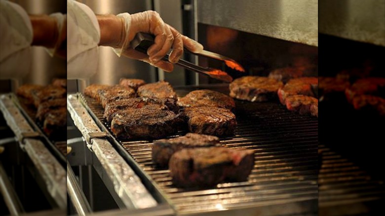 Gloved hand using tongs to arrange steaks on restaurant-grade broiler rack.