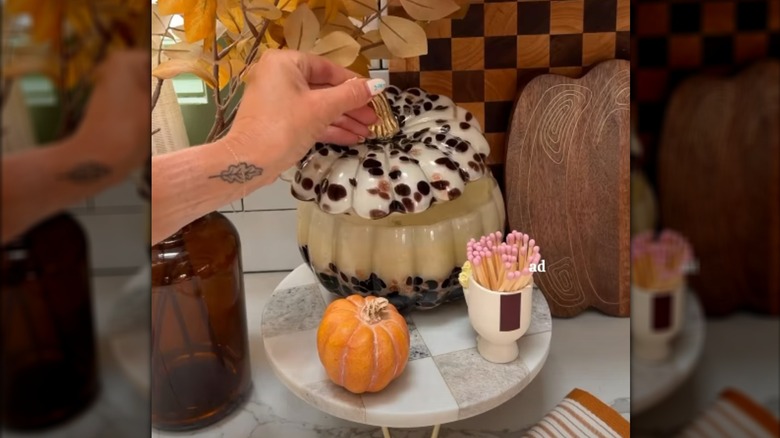 Hand lifting lid from pumpkin shaped candle on pedestal